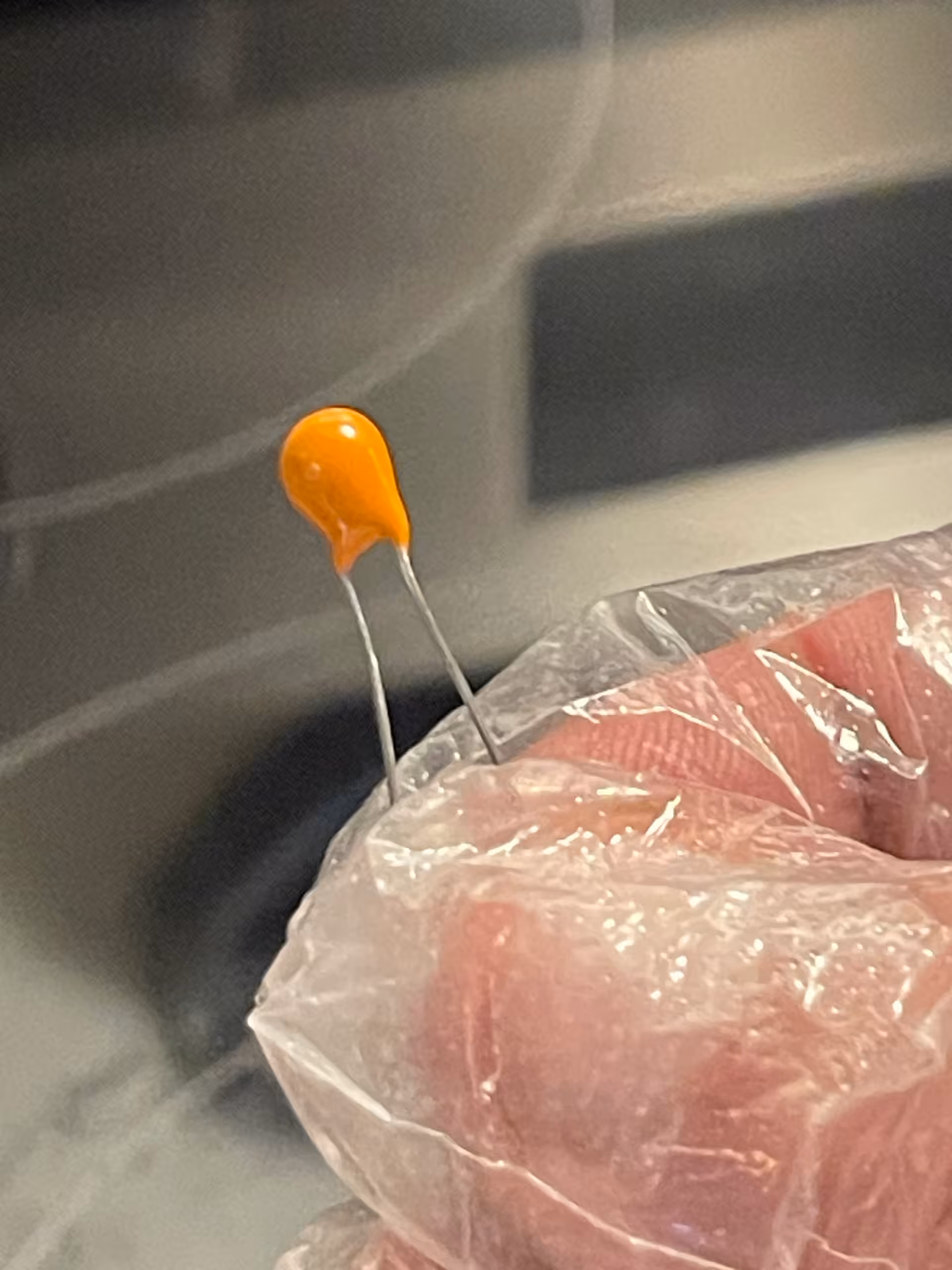 Small orange teardrop tantalum capacitor on two radial leads, held above a gloved hand, unmarked side.