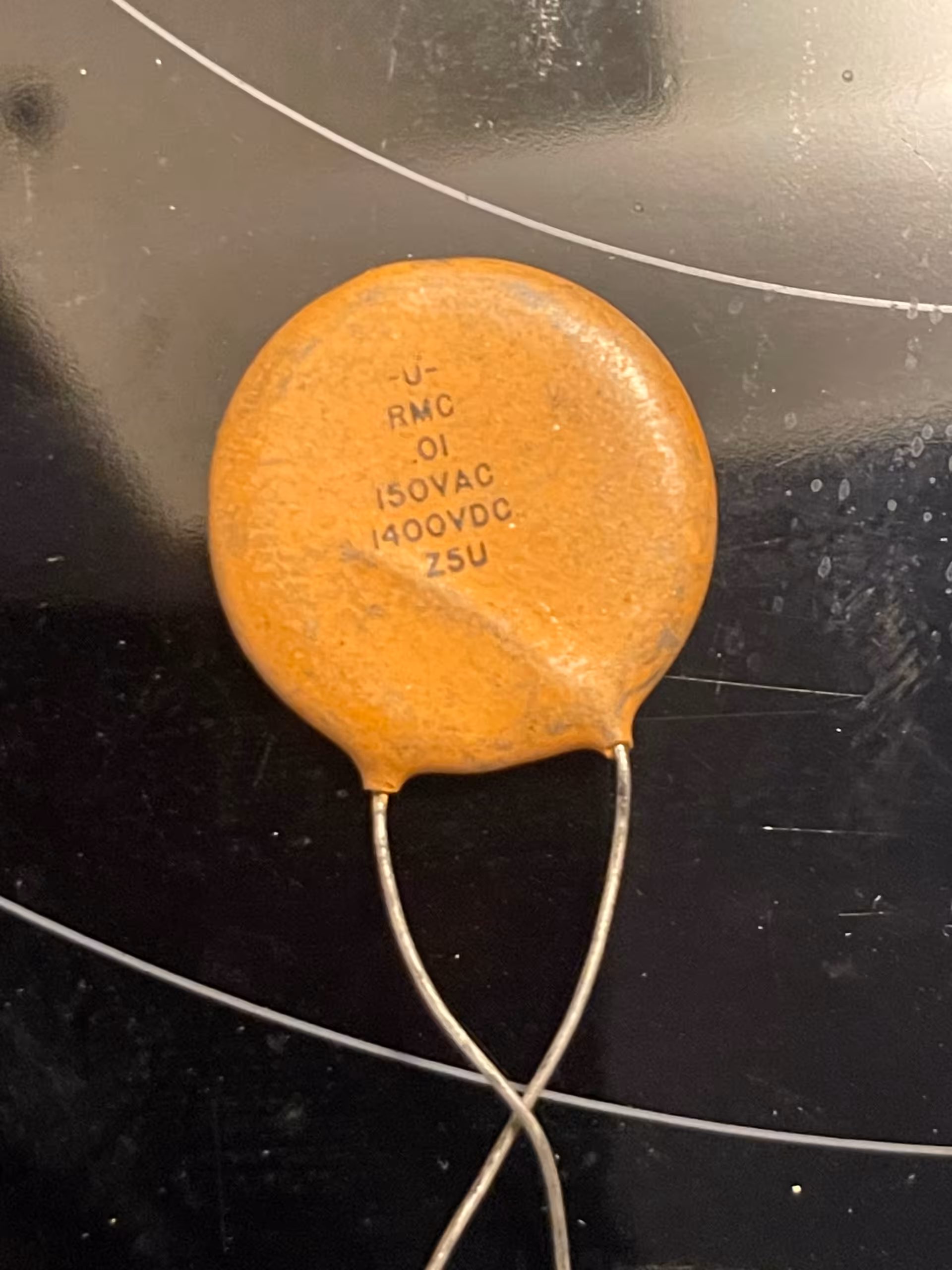 Single orange ceramic disc capacitor on black glass, marked -U- RMC .01 150VAC 1400VDC Z5U.